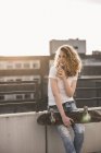 Young woman with skateboard drinking beverage on roof terrace at sunset — Stock Photo