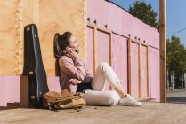 Smiling young woman sitting on platform next to guitar case and listening to music — Stock Photo