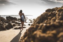 Francia, Bretagna, giovane donna che porta la tavola da surf su una spiaggia rocciosa al mare — Foto stock