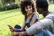Coppia felice seduta sul prato in un parco, utilizzando lo smartphone — Foto stock