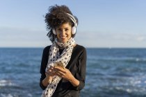 Portrait de femme souriante écoutant de la musique avec smartphone et écouteurs en face de la mer — Photo de stock