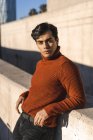 Portrait de jeune homme à la mode portant pull col roulé appuyé sur un mur à la lumière du soleil — Photo de stock