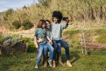 Famiglia felice seduta su una panchina in un giardino, madre che si fa selfie — Foto stock