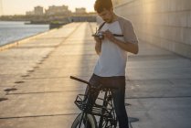 Young man with commuter fixie bike and camera on waterfront promenade at the riverside — Stock Photo