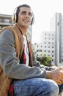 Un jeune homme souriant assis à l'extérieur dans la ville portant des écouteurs — Photo de stock