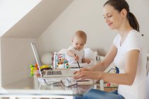 Sorrindo mãe usando laptop e filhinha brincando na mesa em casa — Fotografia de Stock