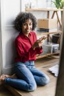 Portrait of smiling woman sitting on the floor at home with cell phone — Stock Photo