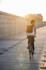 Young man with backpack riding bike on promenade at sunset — Stock Photo