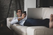 Щасливий молодий чоловік лежить вдома на дивані, користуючись мобільним телефоном і ноутбуком. — стокове фото