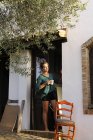 España, Andalucía, mujer joven con taza de café de pie descalzo en la entrada de la casa mirando a la distancia - foto de stock