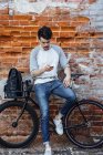 Young man with commuter fixie bike standing at brick wall with cell phone and earphones — Stock Photo