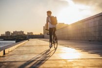 Young man with backpack riding bike on waterfront promenade at the riverside at sunset — Stock Photo