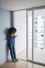 Sorridente giovane donna in piedi alla porta del balcone aperto del suo nuovo appartamento utilizzando lo smartphone — Foto stock