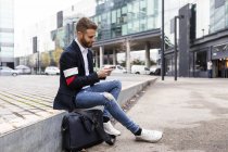 Стильный бизнесмен, сидящий в городе с помощью мобильного телефона — стоковое фото