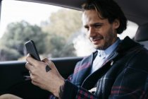 Hombre sonriente sentado en el asiento trasero de un coche usando el teléfono celular - foto de stock