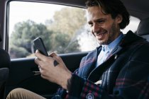 Hombre sonriente sentado en el asiento trasero de un coche usando el teléfono celular - foto de stock