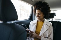 Mulher sorridente sentada no banco de trás de um carro usando telefone celular — Fotografia de Stock
