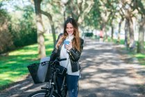 Smiling young woman with bicycle in park using cell phone — Stock Photo