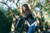 Smiling young woman with bicycle in park using cell phone — Stock Photo