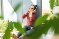 Mujer sonriente con auriculares y portátil sentado en la ventana en casa - foto de stock