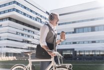 Hombre de negocios maduro sonriente con bicicleta, café para llevar y auriculares sobre la marcha en la ciudad - foto de stock