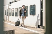 Lächelnder junger Mann mit Kopfhörer, Handy und Koffer am Bahnsteig — Stockfoto