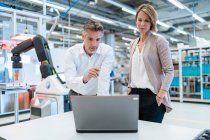 Businessman and businesswoman with laptop talking in a modern factory hall — Stock Photo