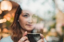 Jeune femme dans un café buvant du café derrière la fenêtre — Photo de stock