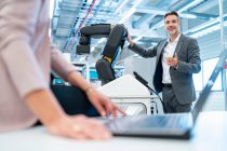 Businessman and businesswoman with laptop and robot in a modern factory hall — Stock Photo