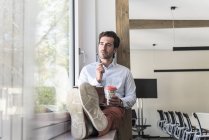 Giovane uomo d'affari seduto sul davanzale della finestra, utilizzando tablet digitale, bere caffè — Foto stock
