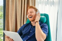 Young man sitting in armchair, listening music, using digital tablet — Stock Photo