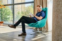Young man sitting in armchair, listening music, using digital tablet — Stock Photo