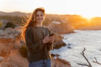 Портрет усмішки почервонілої дівчини з мобільним телефоном біля узбережжя на заході сонця (Івіца, Іспанія). — стокове фото
