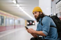 Man with backpack listening music with smartphone and earphones while waiting at platform, Berlin, Germany — Stock Photo