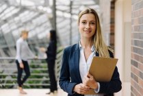 Portrait of a confident young businesswoman in a modern office building with colleagues in background — Stock Photo