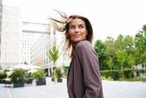 Femme d'affaires souriante aux cheveux balayés par le vent dans la ville se retournant — Photo de stock