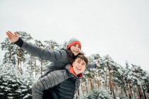 Retrato de un joven sonriente dando a su novia feliz un paseo a cuestas en el bosque de invierno - foto de stock