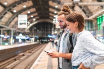 Pareja joven esperando el tren en la estación y usando smartphone, Berlín, Alemania - foto de stock