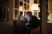 Felice coppia anziana sdraiata sul divano a casa di notte utilizzando il computer portatile — Foto stock