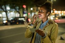 Portrait d'une jeune femme souriante utilisant des écouteurs et un smartphone dans la ville la nuit, Lisbonne, Portugal — Photo de stock