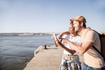 Jeune couple utilisant un téléphone portable sur la jetée au bord de l'eau, Lisbonne, Portugal — Photo de stock