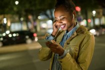 Portrait d'une jeune femme souriante utilisant des écouteurs et un smartphone dans la ville la nuit, Lisbonne, Portugal — Photo de stock