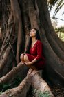 Bella giovane donna che indossa un vestito rosso accovacciato su un albero con grandi radici — Foto stock