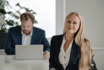 Портрет уверенной деловой женщины в офисе с бизнесменом на заднем плане — стоковое фото