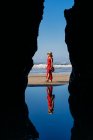Mujer rubia vestida de rojo y sombrero y caminando por la playa, Arco Natural en Playa de Las Catedrales, España - foto de stock