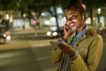 Portrait d'une jeune femme souriante utilisant des écouteurs et un smartphone dans la ville la nuit, Lisbonne, Portugal — Photo de stock