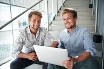 Ritratto di due giovani uomini d'affari sorridenti seduti sulle scale con laptop — Foto stock