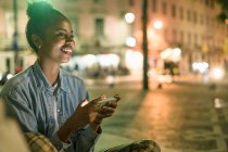 Portrait of happy young woman using smartphone in the city by night, Lisbon, Portugal — Stock Photo