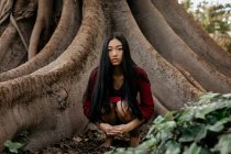 Bella giovane donna che indossa un vestito rosso accovacciato su un albero con grandi radici — Foto stock