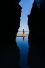 Mujer rubia vestida de rojo y sombrero y caminando por la playa, Arco Natural en Playa de Las Catedrales, España - foto de stock